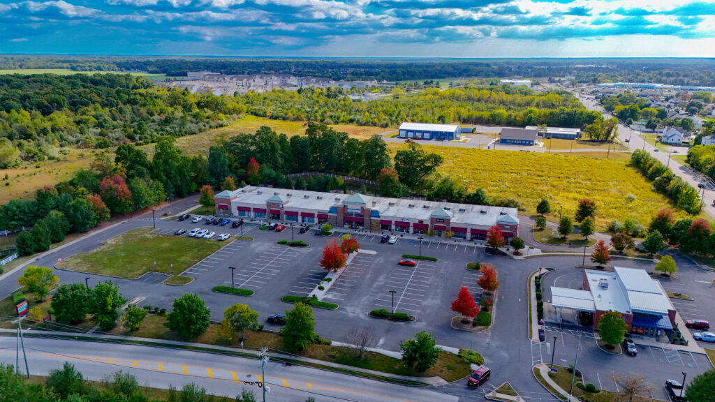 Aerial view of a strip mall site with large warehouses in the background, paved areas, parked vehicles, and surrounding greenery on a clear day.