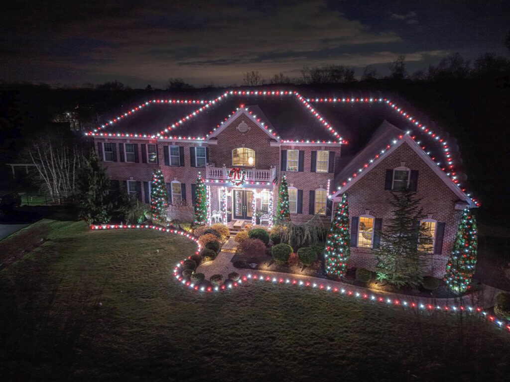 A large brick house decorated with red and white Christmas lights along the roofline, windows, and landscaping, creating a festive nighttime scene.