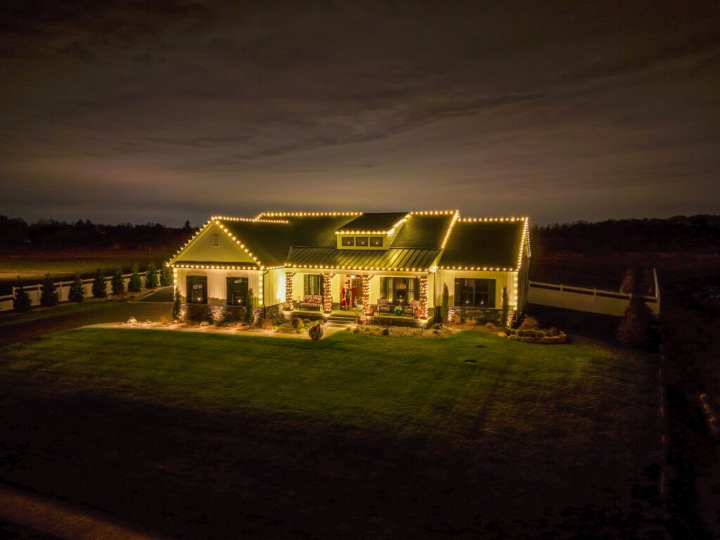 A well-lit modern farmhouse decorated with warm white string lights during the nighttime, surrounded by a neatly landscaped lawn and a fence in the background.